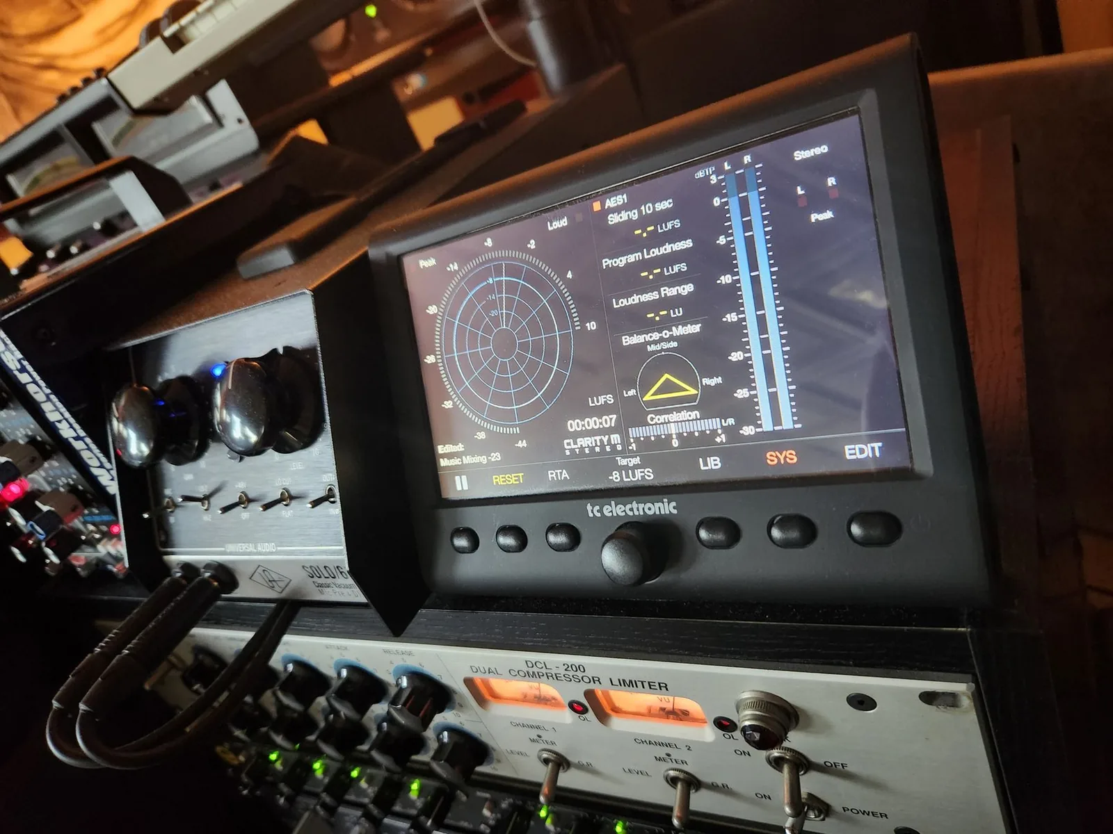 Photo of a TC Electronic Clarity M hardware meter displaying LUFS integrated loudness, true peak, and loudness range readings on its front-panel LCD.